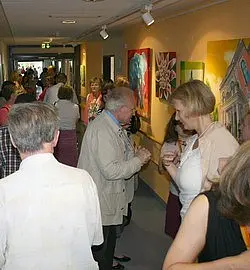 Ein belebter Flur mit vielen Personen, die sich unterhalten und eine Kunstausstellung besuchen. An den Wänden hängen verschiedene bunte Bilder. Die Atmosphäre wirkt gesellig und einladend.