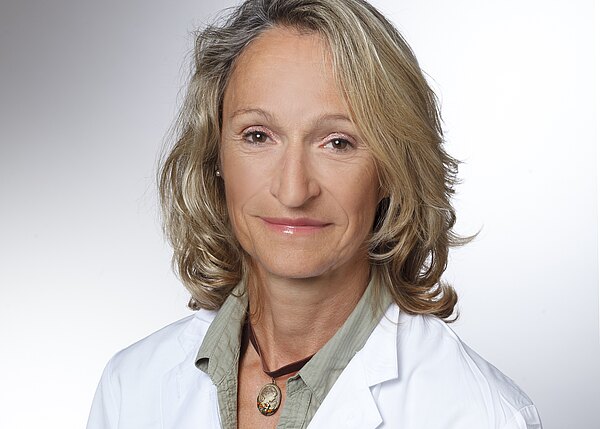 Porträt einer erwachsenen Frau in weißem Arztkittel, leichtes Lächeln vor neutralem Hintergrund; wirkt professionell und freundlich.