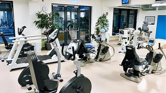 Heller, leerer Fitnessraum. Mehrere Cardio- und Rehageräte stehen geordnet: Ergometer, Liegeräder, Laufband und weitere Trainingsmaschinen. Moderne, saubere Umgebung.