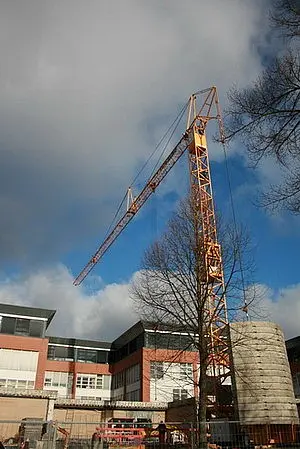 Ein massiver Kran steht neben einem Gebäude. Der Kran hebt einen großen zylinderförmigen Betonblock an. Der Himmel ist bewölkt, mit einigen blauen Stellen sichtbar. Die Umgebung ist Baustelle mit Baugeräten und einem gesperrten Bereich.