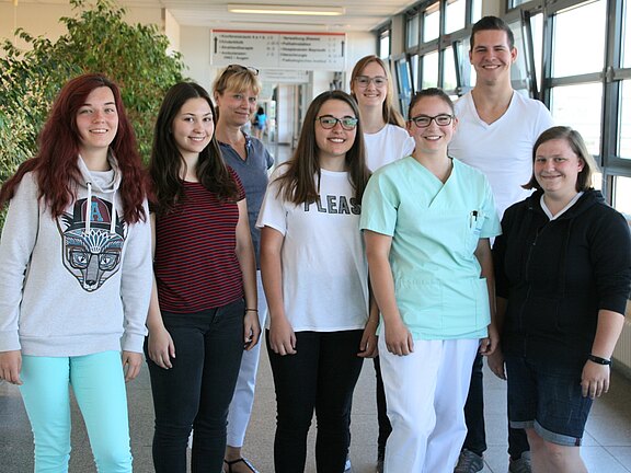 Gruppenfoto in einem hellen Flur: acht junge Personen stehen nebeneinander und lächeln in die Kamera. Die meisten tragen Freizeitkleidung, zwei Personen tragen weiße bzw. mintgrüne Berufskleidung, was auf ein medizinisches Umfeld hindeutet.