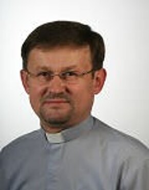 Das Bild zeigt einen Mann in einem grauen Hemd mit einer weißen Kragen. Er hat kurze, dunkle Haare und trägt eine Brille. Sein Gesichtsausdruck ist freundlich und ernst. Der Hintergrund ist hell und einfarbig.