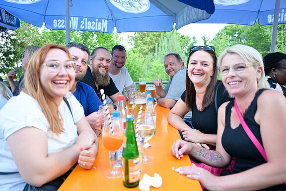 Acht Erwachsene sitzen an einem orangefarbenen Tisch im Freien, lachen in die Kamera und trinken Bier und Limonade. Gesellige, sommerliche Biergartenstimmung.