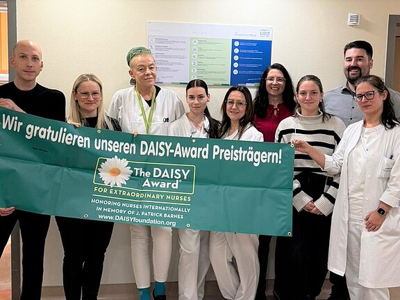 Gruppe von Pflegekräften und Kolleginnen/Kollegen im Krankenhausflur hält ein großes grünes Banner mit der Aufschrift: „Wir gratulieren unseren DAISY-Award Preisträgern! The DAISY Award for Extraordinary Nurses.“ Feierliche Anerkennung für besondere Pflegeleistungen.