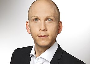 Christian Brodmerkel Ein junger Mann mit kurzem, glattem Kopf trägt einen schwarzen Anzug und ein weißes Hemd. Er schaut direkt in die Kamera mit neutrale Miene. Der Hintergrund ist hell und unscharf, wodurch der Fokus auf seinem Gesicht liegt.