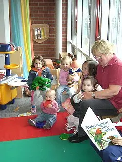 Eine Gruppe von sechs Kindern sitzt auf einem bunten Teppich, während eine Erwachsene sie mit einem Buch anspricht. Einige Kinder halten Spielzeug in den Händen. Die Umgebung wirkt freundlich und kindgerecht.