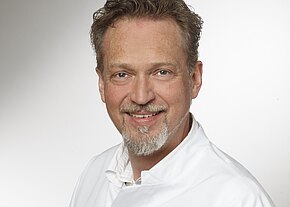 Porträt eines lächelnden Mannes mittleren Alters mit kurzem lockigem Haar und grauem Kinnbart. Er trägt einen weißen Kittel; neutraler Hintergrund.