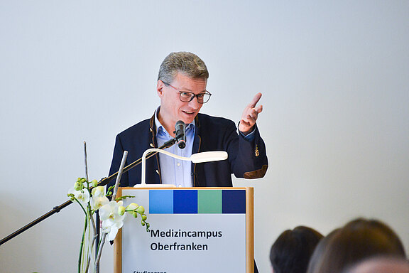 Redner mit Brille hält einen Vortrag am Podium mit der Aufschrift „Medizincampus Oberfranken“ und gestikuliert; Mikrofon und Leselampe sichtbar.