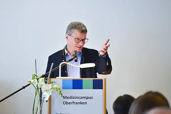 Redner mit Brille hält einen Vortrag am Podium mit der Aufschrift „Medizincampus Oberfranken“ und gestikuliert; Mikrofon und Leselampe sichtbar.