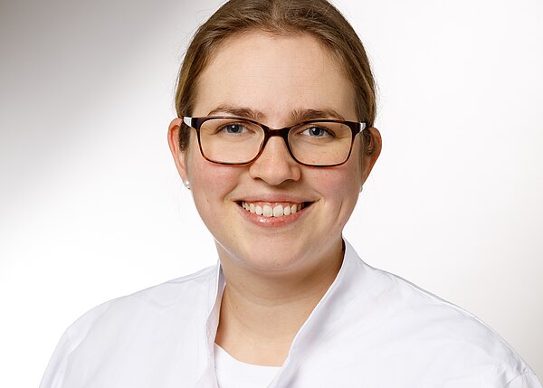 Porträt einer lächelnden Person mit Brille im weißen Kittel vor hellem, neutralem Hintergrund.