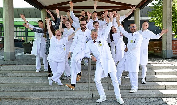 Eine Gruppe von 14 Personen in weißen Kitteln steht auf Treppen mit gehobenen Händen und lächelt. Sie zeigen Freude und Begeisterung in einer freundlichen, einladenden Umgebung.