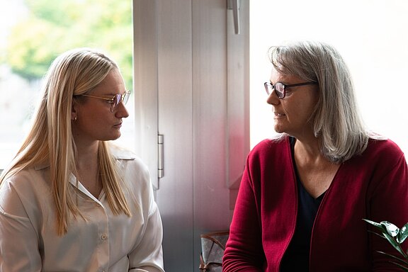 Zwei Frauen sitzen nebeneinander vor einem hellen Fenster und sprechen miteinander. Die Jüngere trägt eine weiße Bluse, die Ältere eine rote Strickjacke; beide haben Brillen. Der Fokus liegt auf ihrem ruhigen Gespräch in entspannter, natürlicher Umgebung.