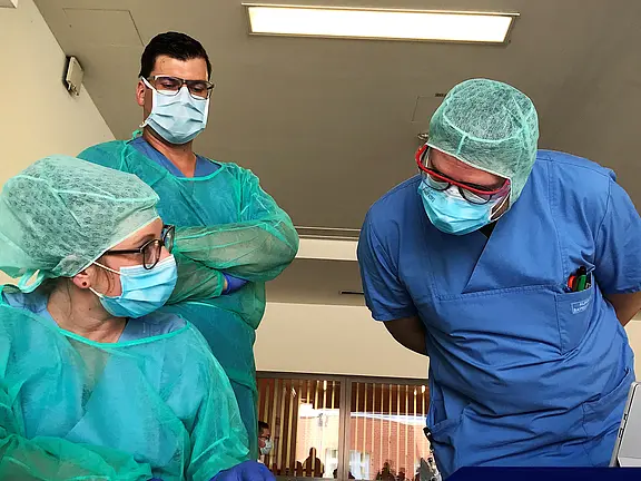 Im Bild sind drei medizinische Fachkräfte in Schutzausrüstung zu sehen. Zwei Personen stehen und schauen auf eine dritte Person, die sitzt und etwas untersucht. Alle tragen Masken und Schutzbrillen. Die Umgebung wirkt klinisch und professionell.