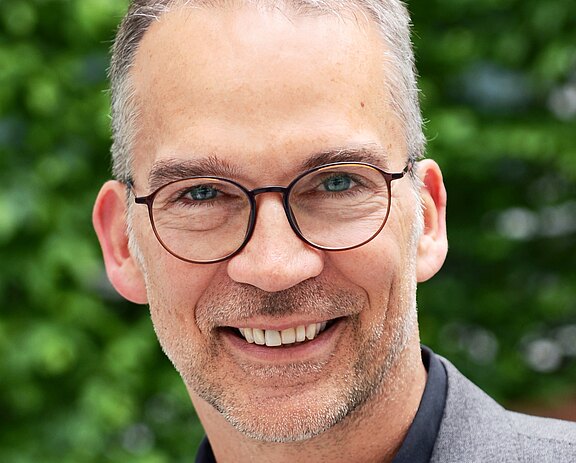 Porträt einer lächelnden Person mit runder Brille und leichtem Bart, die direkt in die Kamera blickt. Kurzes, grau meliertes Haar, dunkles Jackett. Hintergrund unscharf mit grünem Laub.