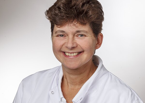 Porträt einer lächelnden erwachsenen Person mit kurzem Haar in weißem Arztkittel vor neutralem Hintergrund; Kopf-und-Schultern-Aufnahme.