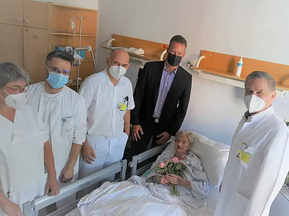 Ein älterer Mensch liegt in einem Krankenhausbett und hält einen Blumenstrauß. Um sie herum stehen fünf Personen, darunter medizinisches Personal und ein Mann in Anzug. Alle tragen Masken. Die Atmosphäre ist freundlich und zeigt eine Unterstützungserfahrung im Krankenhaus.
