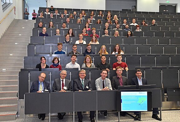Gruppenfoto in einem großen Hörsaal: Viele Studierende sitzen in Reihen und schauen zur Kamera. In der ersten Reihe sitzen mehrere Personen an einem Tisch mit Bildschirm, der eine Präsentationsfolie zeigt. Der Saal ist hell und modern, die Stimmung wirkt formell.