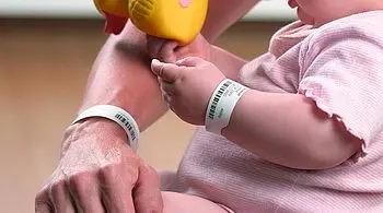 Ein Elternteil hält eine gelbe Quietscheente, während ein Baby in rosa Kleidung spielt. Beide tragen Armbänder, vermutlich zur Identifikation. Der Fokus liegt auf der Interaktion zwischen dem Baby und dem Spielzeug.