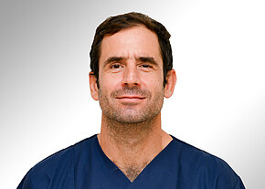 Porträt eines mittelalten Mannes mit kurzem dunklem Haar, leichtem Bart und neutralem Lächeln. Er trägt dunkelblaue OP-/Pflegekleidung. Kopf und Oberkörper frontal vor hellem, neutralem Hintergrund.