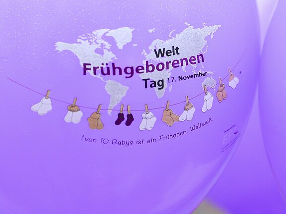 Lila Ballon mit Aufdruck zum Weltfrühgeborenentag (17. November): Vor einer Weltkarte hängen kleine Babysöckchen an einer Leine. Begleittext: „1 von 10 Babys ist ein Frühchen. Weltweit.“