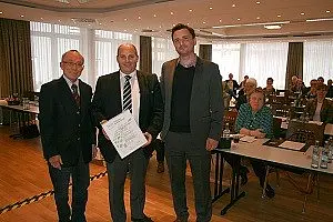 Drei Männer stehen in einem Konferenzraum zusammen. Der Mann in der Mitte hält ein Dokument in den Händen. Im Hintergrund sind Tische mit Teilnehmern, die an einer Besprechung teilnehmen. Die Atmosphäre wirkt formell und professionell.