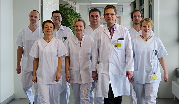 Eine Gruppe von sieben medizinischen Fachkräften in weißen Kitteln steht in einem Krankenhausflur. Sie schauen freundlich in die Kamera und wirken professionell. Die Umgebung ist hell erleuchtet, mit einigen Pflanzen im Hintergrund.