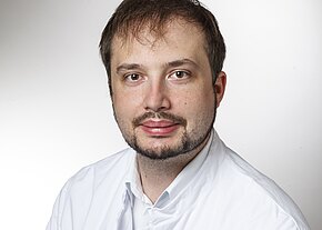 Dr. Torsten Kämpf Porträt eines erwachsenen Mannes mit kurzem braunen Haar und Bart, leicht lächelnd. Er trägt einen weißen Kittel und blickt direkt in die Kamera vor hellem Hintergrund.