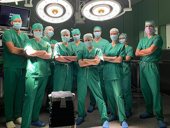 Eine Gruppe von zehn Fachleuten im Operationssaal steht in grüner Arbeitskleidung und Masken. Sie posieren entschlossen vor einem chirurgischen Tisch, umgeben von medizinischen Geräten und OP-Lichtern. Die Atmosphäre wirkt professionell und konzentriert.