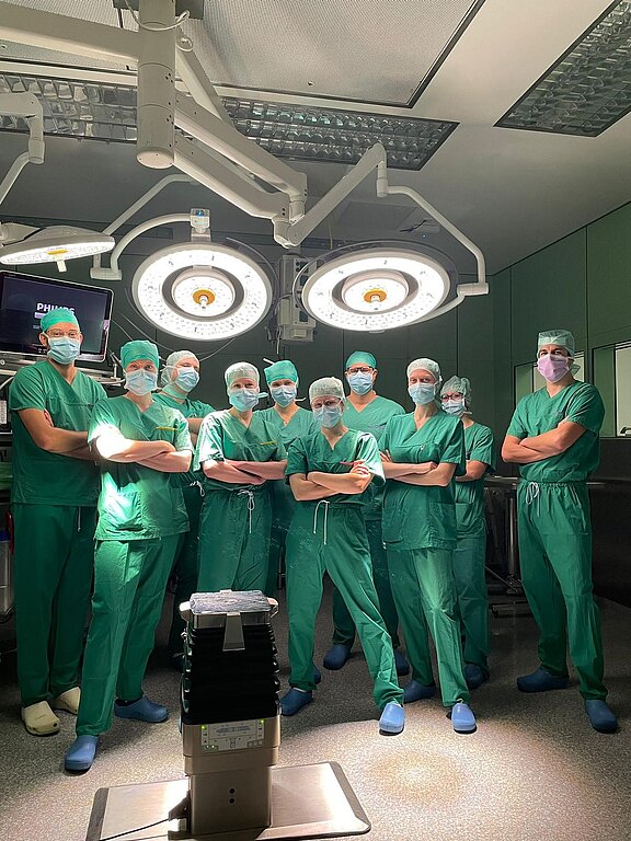 Eine Gruppe von zehn Fachleuten im Operationssaal steht in grüner Arbeitskleidung und Masken. Sie posieren entschlossen vor einem chirurgischen Tisch, umgeben von medizinischen Geräten und OP-Lichtern. Die Atmosphäre wirkt professionell und konzentriert.