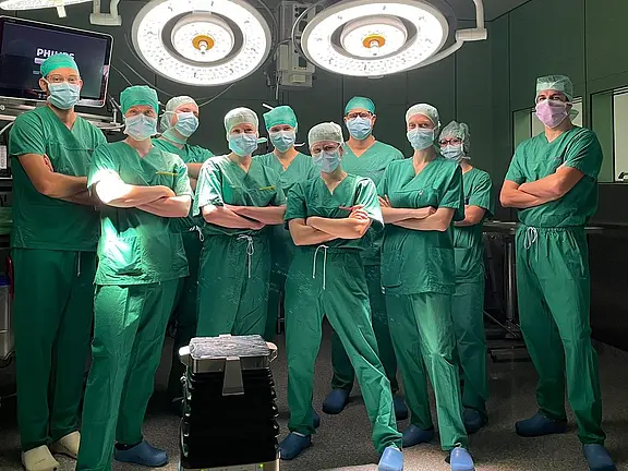 Eine Gruppe von zehn Fachleuten im Operationssaal steht in grüner Arbeitskleidung und Masken. Sie posieren entschlossen vor einem chirurgischen Tisch, umgeben von medizinischen Geräten und OP-Lichtern. Die Atmosphäre wirkt professionell und konzentriert.