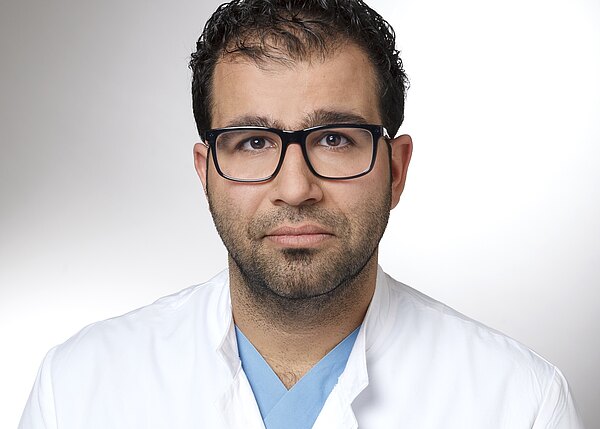 Portrait einer Person mit dunklem Haar und schwarzer Brille, in weißem Arztkittel über blauer OP-Kleidung. Blick direkt zur Kamera, ruhiger Ausdruck, heller neutraler Hintergrund.