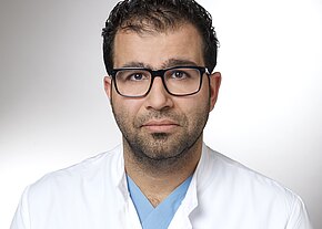 Portrait einer Person mit dunklem Haar und schwarzer Brille, in weißem Arztkittel über blauer OP-Kleidung. Blick direkt zur Kamera, ruhiger Ausdruck, heller neutraler Hintergrund.