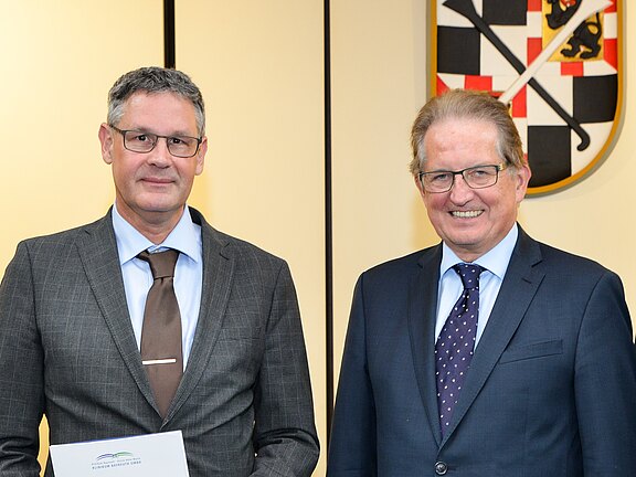 Drei Männer in Anzügen stehen nebeneinander und lächeln in die Kamera. Die beiden außen halten offizielle Dokumente. Im Hintergrund hängt ein Wappen. Szene einer formellen Übergabe oder Ehrung in einem Behördensaal.