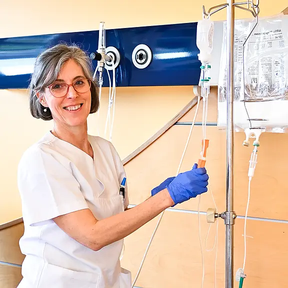 Eine Krankenschwester mit grauen Haaren und Brille lächelt, während sie an einer Infusion arbeitet. Sie trägt Handschuhe und hält ein Werkzeug in der Hand, um einen IV-Zugang zu kontrollieren. Im Hintergrund ist ein Pflegebett und medizinische Ausrüstung zu sehen.