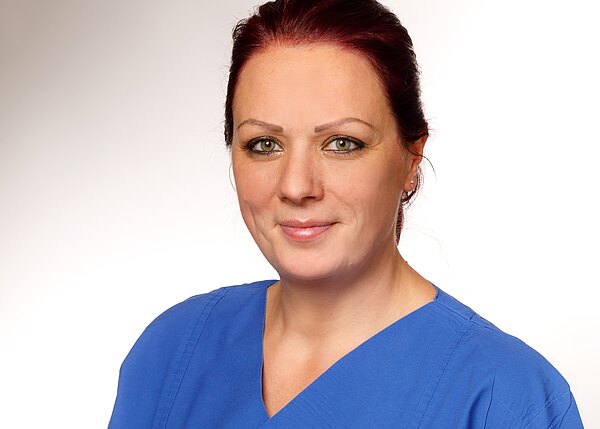 Porträt einer lächelnden Frau mit kurzen, roten Haaren, die ein blaues medizinisches Hemd trägt. Der Hintergrund ist hell und neutral, was die Aufmerksamkeit auf ihr Gesicht lenkt. Sie hat grüne Augen und wirkt freundlich und professionell.