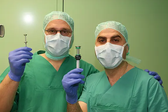 Zwei medizinische Fachkräfte in grüner OP-Kleidung, Masken und Handschuhen stehen nebeneinander und halten eine Spritze bzw. ein Injektionsgerät in die Kamera. Die Szene wirkt wie eine Vorbereitung auf eine medizinische Behandlung oder Impfung.