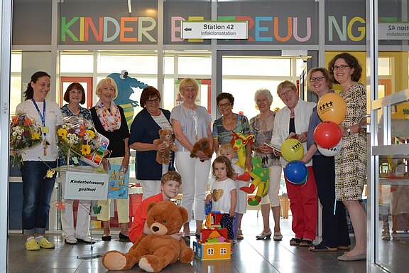 Gruppe von Erwachsenen und zwei Kindern in einem Krankenhausflur, vor einer Tür mit „Kinderbetreuung“. Sie halten bunte Luftballons, Blumen und Spielzeug; große Stofftiere und Bauklötze vorn. Feierliche Szene, wahrscheinlich zur Eröffnung der Kinderbetreuung.