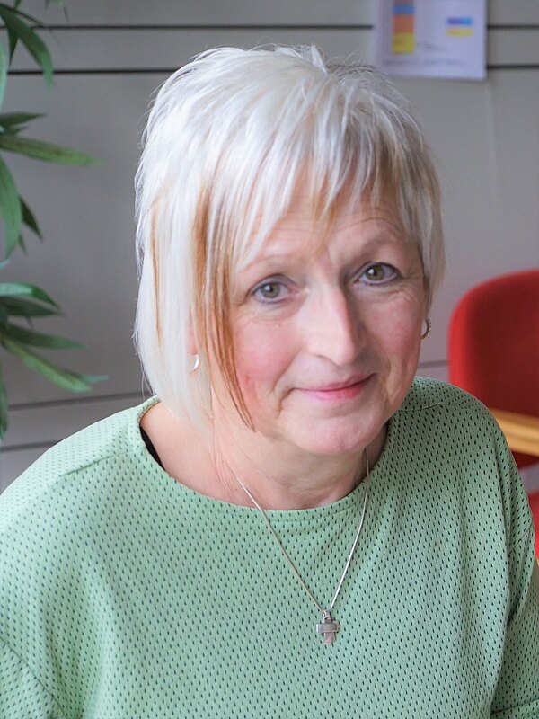 Nahaufnahme einer Frau mit kurzem weißblondem Haar. Sie trägt ein grünes Oberteil und eine silberne Kette und blickt freundlich in die Kamera. Innenraum mit schlichtem, neutralem Hintergrund.