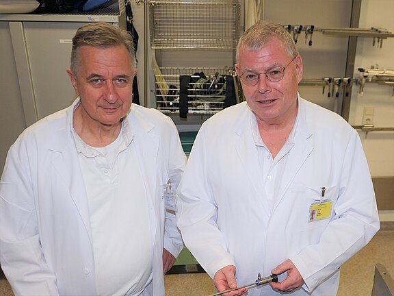 Zwei Mediziner in weißen Kitteln in einem Klinikraum; der rechte hält ein langes chirurgisches Instrument, vermutlich ein Endoskop. Beide schauen in die Kamera.