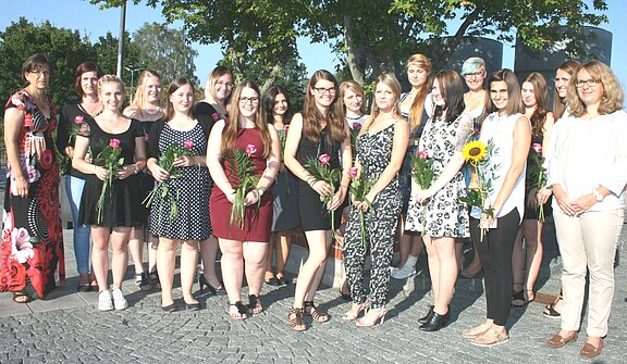 Gruppenporträt von jungen Frauen, die mit Blumen in der Hand an einer Feier teilnehmen. Sie tragen verschiedene festliche Outfits und lächeln. Ein paar Lehrkräfte stehen ebenfalls dabei. Im Hintergrund sind Bäume und ein klarer Himmel sichtbar.