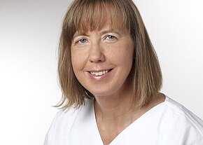 Station 2a/2b Porträt einer lächelnden Frau mit schulterlangem hellbraunem Haar und Pony, in weißer Kleidung vor neutralem, hellem Hintergrund.