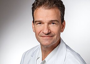Dr. med. Rainer Krauß Porträt eines freundlich blickenden Mannes mittleren Alters mit kurzen braunen Haaren. Er trägt weiße Kleidung und schaut direkt in die Kamera, vor neutralem Hintergrund.