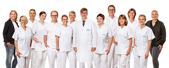 Gruppenfoto eines medizinischen Teams: Mehrere Pflegekräfte in weißen Uniformen und ein Arzt in der Mitte mit Kittel und Krawatte. Alle stehen nebeneinander vor weißem Hintergrund und lächeln freundlich.