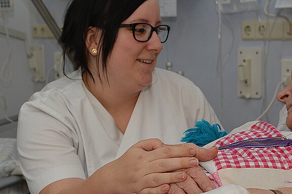 Pflegekraft in weißer Kleidung lächelt und hält die Hand einer im Krankenhausbett liegenden Person; eine beruhigende, fürsorgliche Szene.