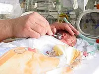 Ein Neugeborenes liegt in einer Krankenhausumgebung. Eine Hand hält ein kleines medizinisches Instrument und untersucht das Baby. Das Baby ist in eine Decke gewickelt und umgeben von medizinischer Ausrüstung.