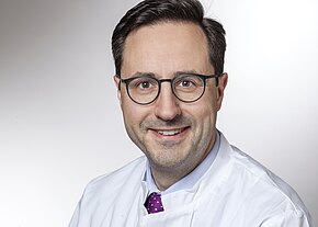 Univ.-Prof. Dr. med. Thorsten Klink, EBCR, EBIR Porträtfoto einer Person mit dunklem Haar und Brille, in weißem Kittel und violetter Krawatte. Sie lächelt und blickt direkt in die Kamera; neutraler, heller Hintergrund.