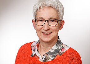 Porträt einer lächelnden erwachsenen Person mit kurzem grauen Haar und dunkler Brille, vor neutralem Hintergrund. Sie trägt ein bunt gemustertes Hemd unter einem orangefarbenen Strickpullover, Blick zur Kamera.