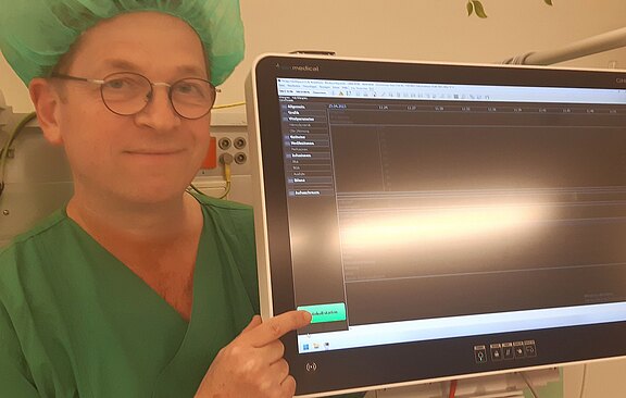 Ein Arzt in grüner Kleidung mit einer Haube lächelt und zeigt auf einen Monitor. Der Bildschirm zeigt überwiegend leere Bereiche mit einigen aufgelisteten Menüpunkten. Er scheint eine Funktion zur Datenanzeige oder -bearbeitung auszuwählen.