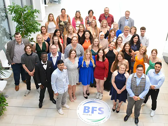 Gruppenfoto von 50 Personen, die in festlicher Kleidung zusammenstehen. Die Gruppe besteht aus Männern und Frauen unterschiedlichen Alters, auf einem hellen Hintergrund mit Pflanzen. Im Vordergrund ist ein Logo mit der Aufschrift "BFS Pflege" zu sehen.