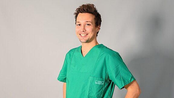 Junger medizinischer Mitarbeiter in grüner OP-Kleidung steht seitlich, lächelt, eine Hand in der Hüfte, vor neutralem Hintergrund.